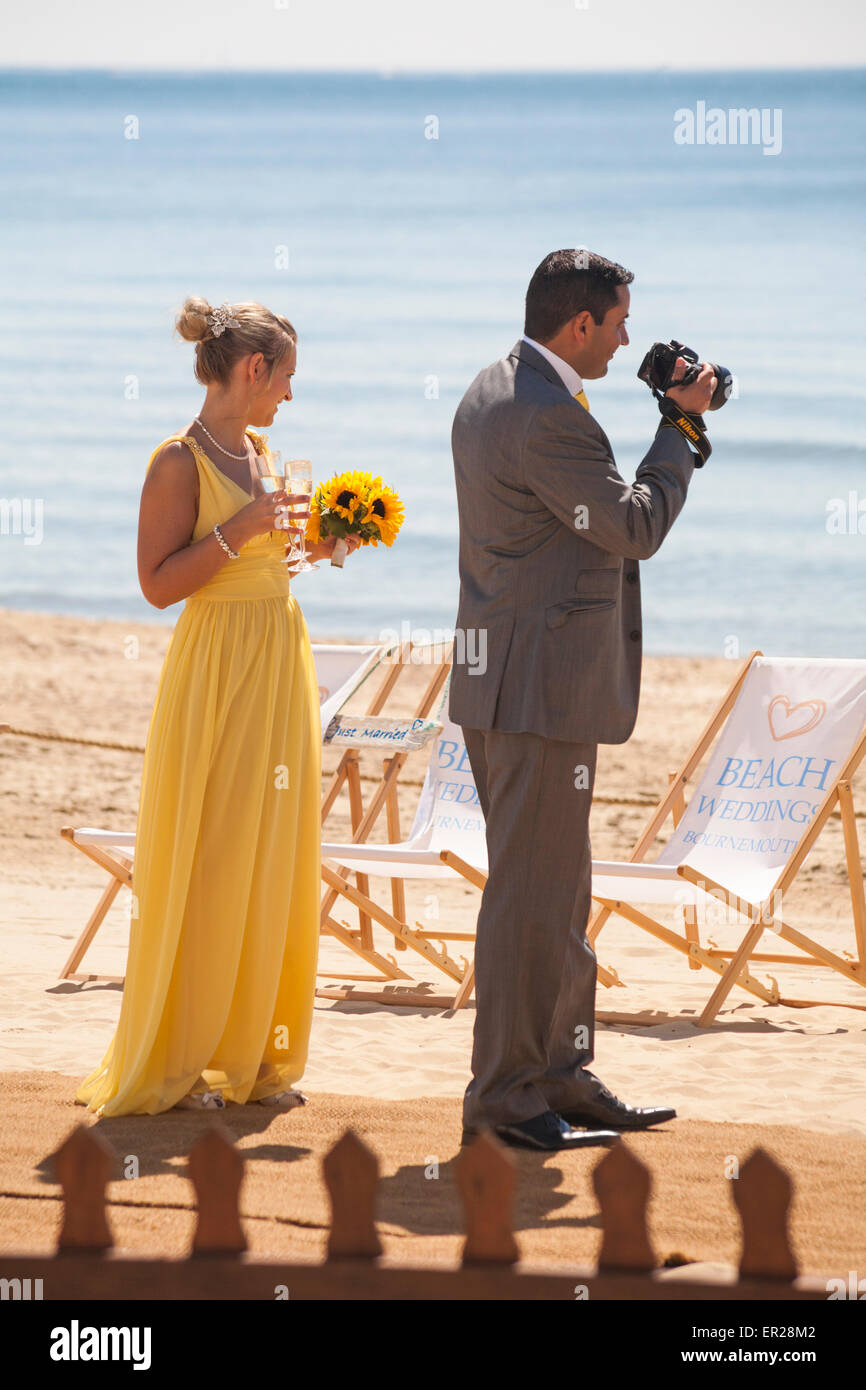 En robe jaune longue de demoiselle d'holding bouquet de tournesols et l'homme avec l'appareil photo lors de la cérémonie du mariage sur la plage de Bournemouth, Mai Banque D'Images