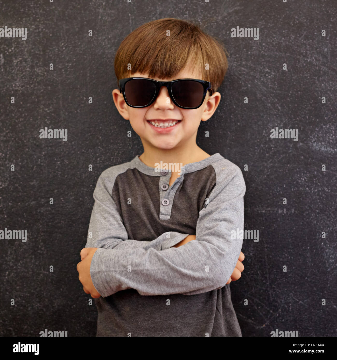 Photo de l'adorable jeune happy boy looking at camera avec lunettes de soleil. Petit enfant souriant, les bras croisés contre le tableau noir Banque D'Images