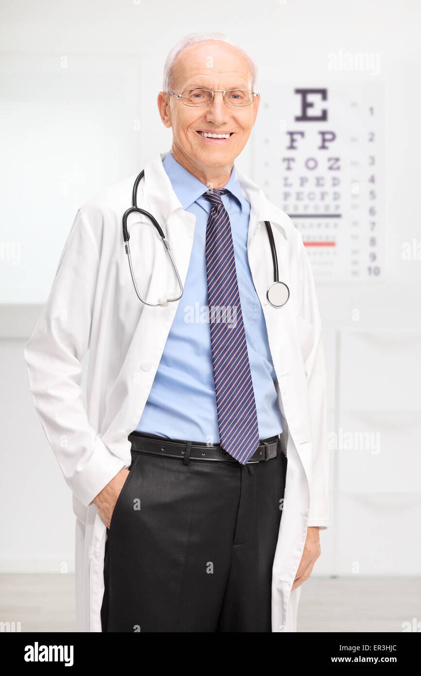 Shot vertical d'un opticien à un manteau blanc posant dans son bureau en face d'un test de la vue et souriant Banque D'Images