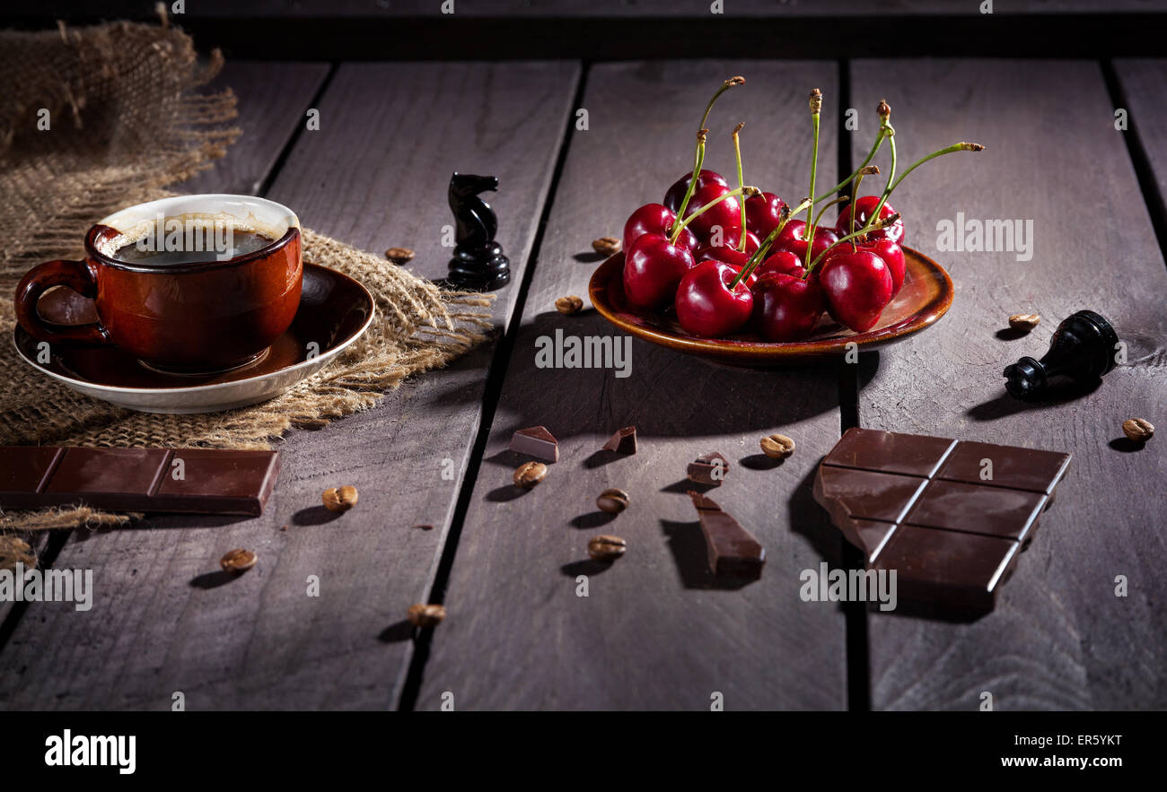Tasse à café, chocolat noir et cerises sur la table en bois avec black chess figure à proximité Banque D'Images