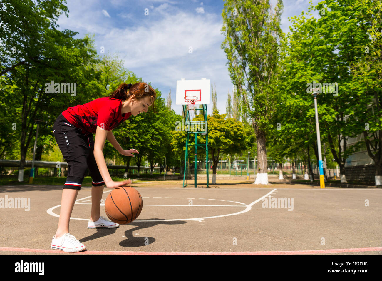 Toute la longueur de jeune femme athlétique Dribbling Basketball à mi Cour dans le parc verdoyant avec panneau et panier en arrière-plan Banque D'Images