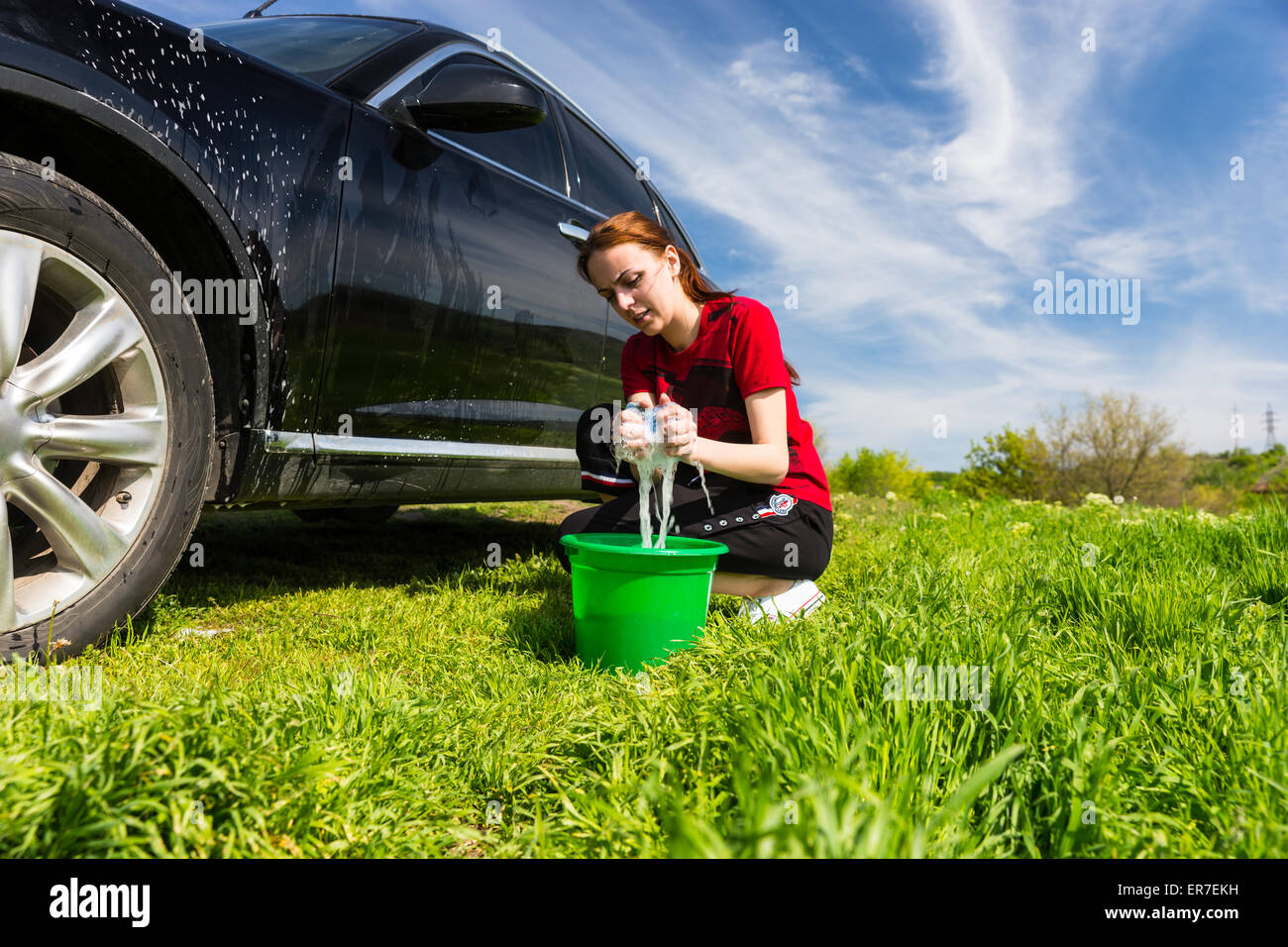 Woman Wearing Red T-Shirt Noir Lavage véhicule en champ, accroupi à côté de la benne verte essorage de l'Éponge savonneuse sur Bright Sunny D Banque D'Images