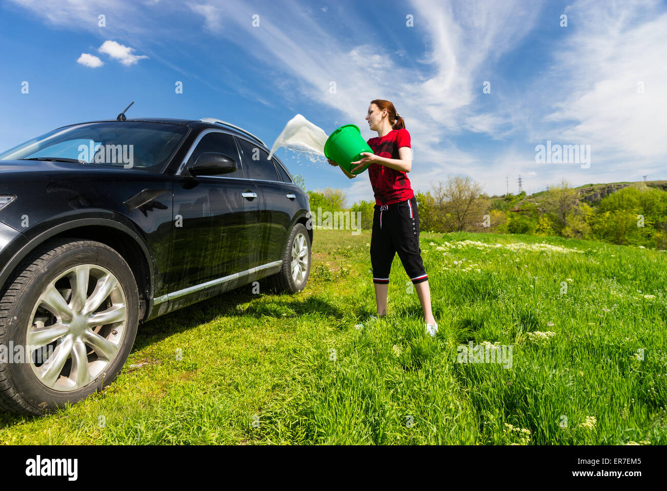 Jeune femme lave-véhicule de luxe Noir à Grassy Green Field sur journée ensoleillée avec ciel bleu, jetant en seau d'eau sur S Banque D'Images