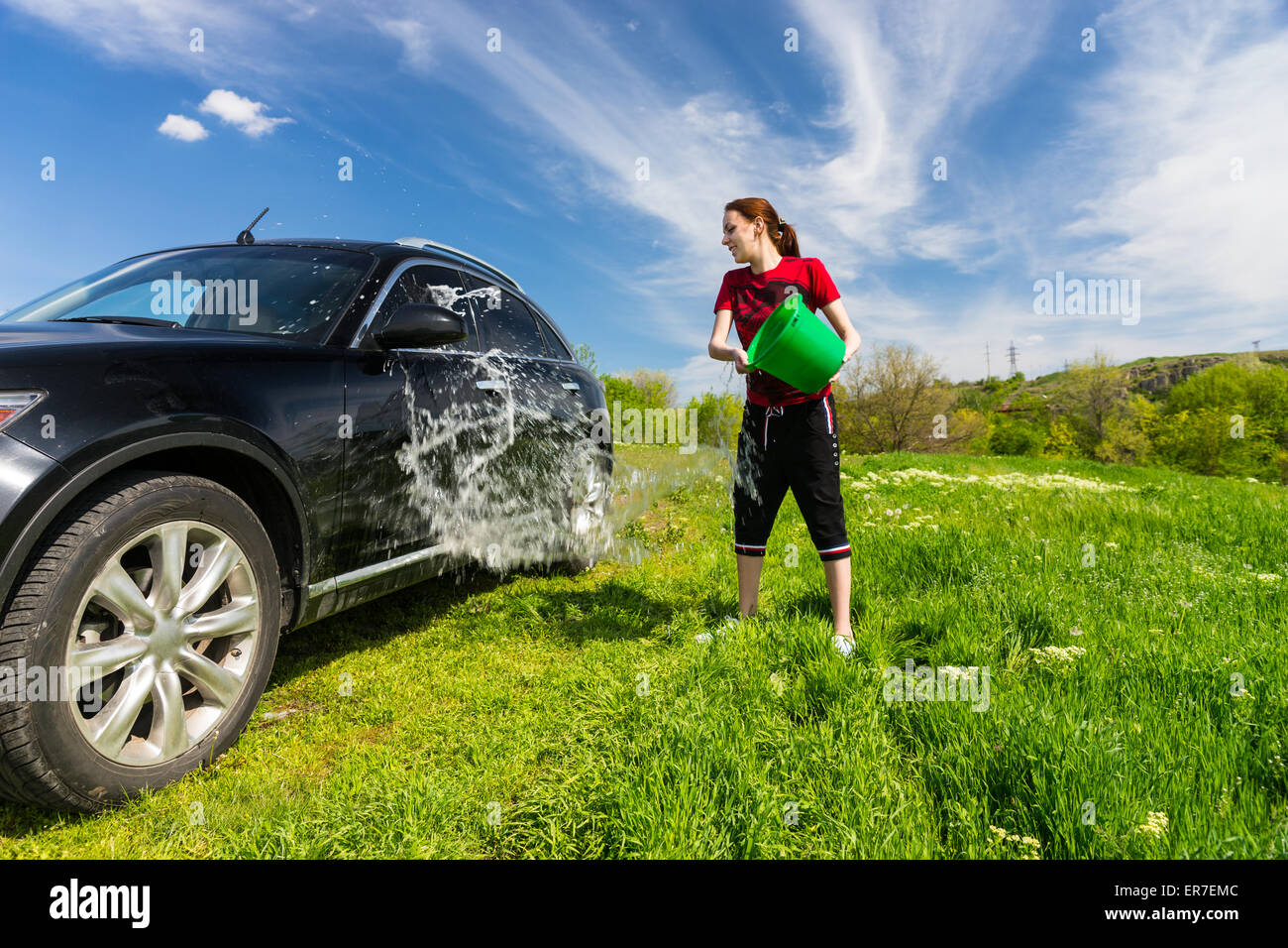 Jeune femme lave-véhicule de luxe Noir à Grassy Green Field sur journée ensoleillée avec ciel bleu, jetant en seau d'eau sur le côté du véhicule. Banque D'Images
