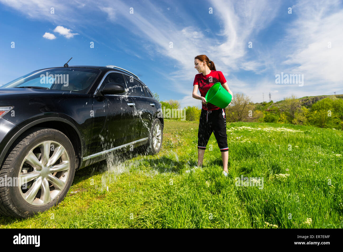 Jeune femme lave-véhicule de luxe Noir à Grassy Green Field sur journée ensoleillée avec ciel bleu, jetant en seau d'eau sur le côté du véhicule. Banque D'Images