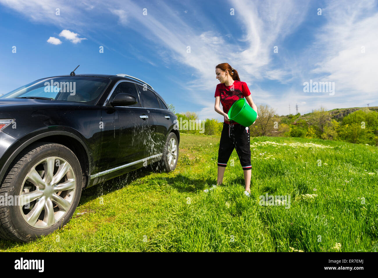 Jeune femme lave-véhicule de luxe Noir à Grassy Green Field sur journée ensoleillée avec ciel bleu, jetant en seau d'eau sur S Banque D'Images