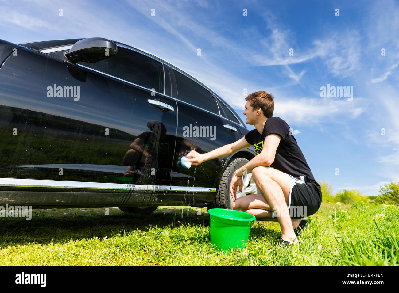 Man voiture avec une éponge savonneuse, accroupi à côté de la benne verte dans les champs verts sur journée ensoleillée avec ciel bleu Banque D'Images