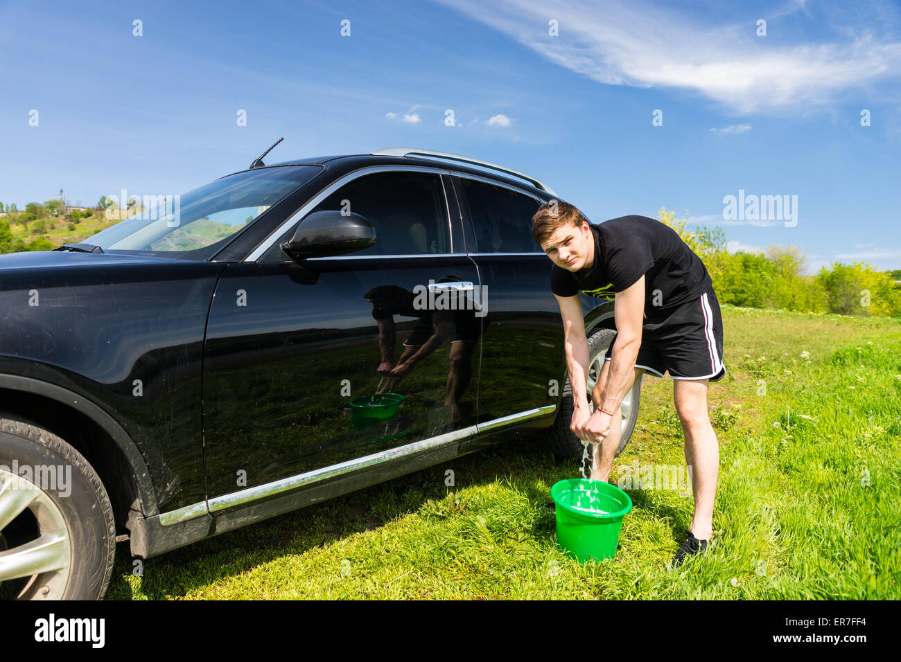 L'homme à la caméra et à l'essorage de l'Éponge savonneuse dans la benne verte pendant le lavage de véhicules de luxe noir en vert sur le terrain herbeux journée ensoleillée avec ciel bleu. Banque D'Images