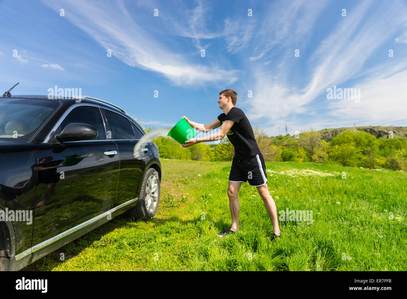Jeune homme noir Lavage véhicule de luxe à Grassy Green Field sur journée ensoleillée avec ciel bleu, jetant en seau d'eau sur le côté du véhicule. Banque D'Images