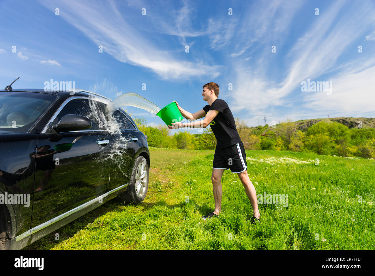 Jeune homme noir Lavage véhicule de luxe à Grassy Green Field sur journée ensoleillée avec ciel bleu, jetant en seau d'eau sur Sid Banque D'Images
