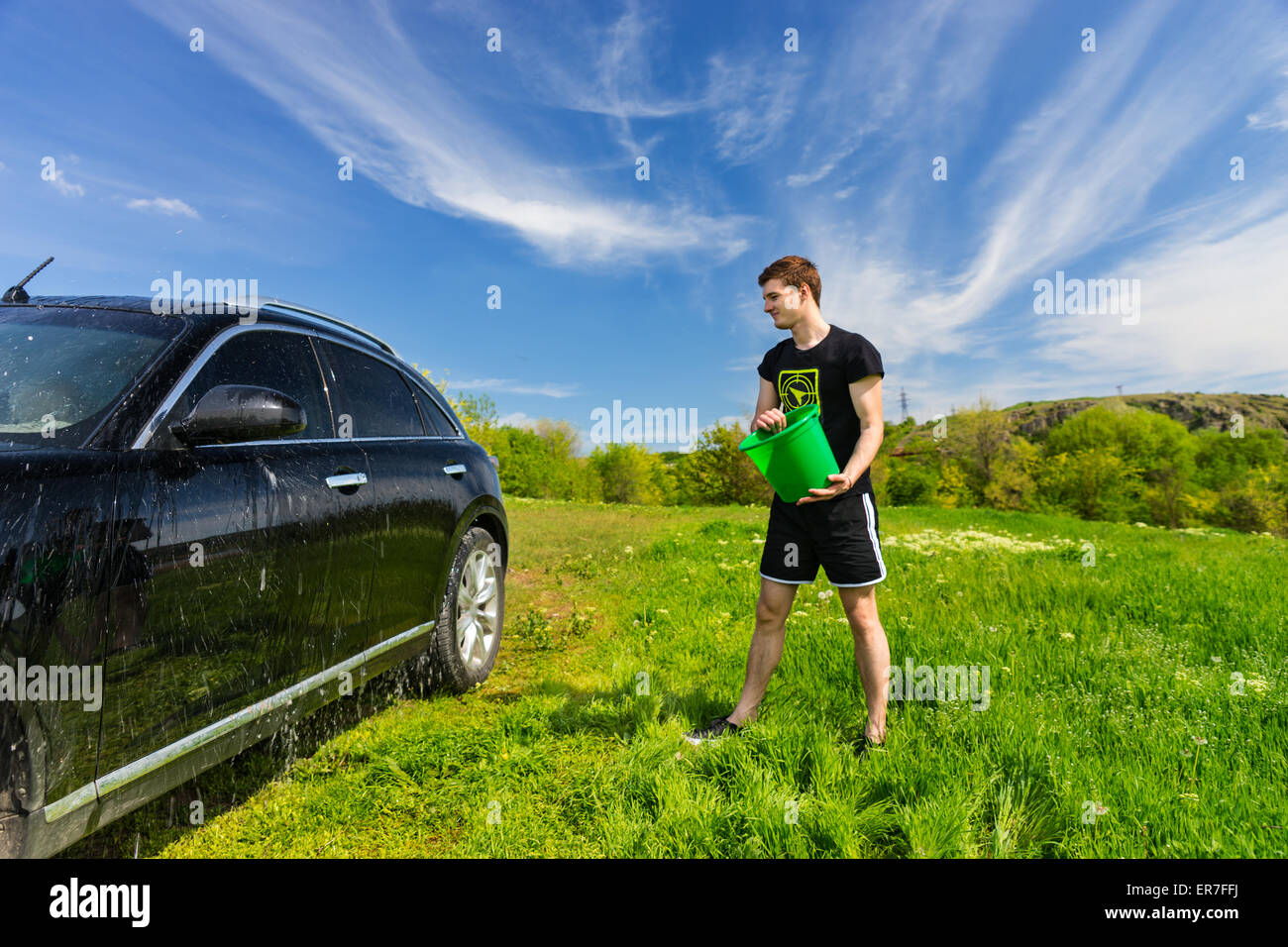 Jeune homme noir Lavage véhicule de luxe à Grassy Green Field sur journée ensoleillée avec ciel bleu, jetant en seau d'eau sur le côté du véhicule. Banque D'Images