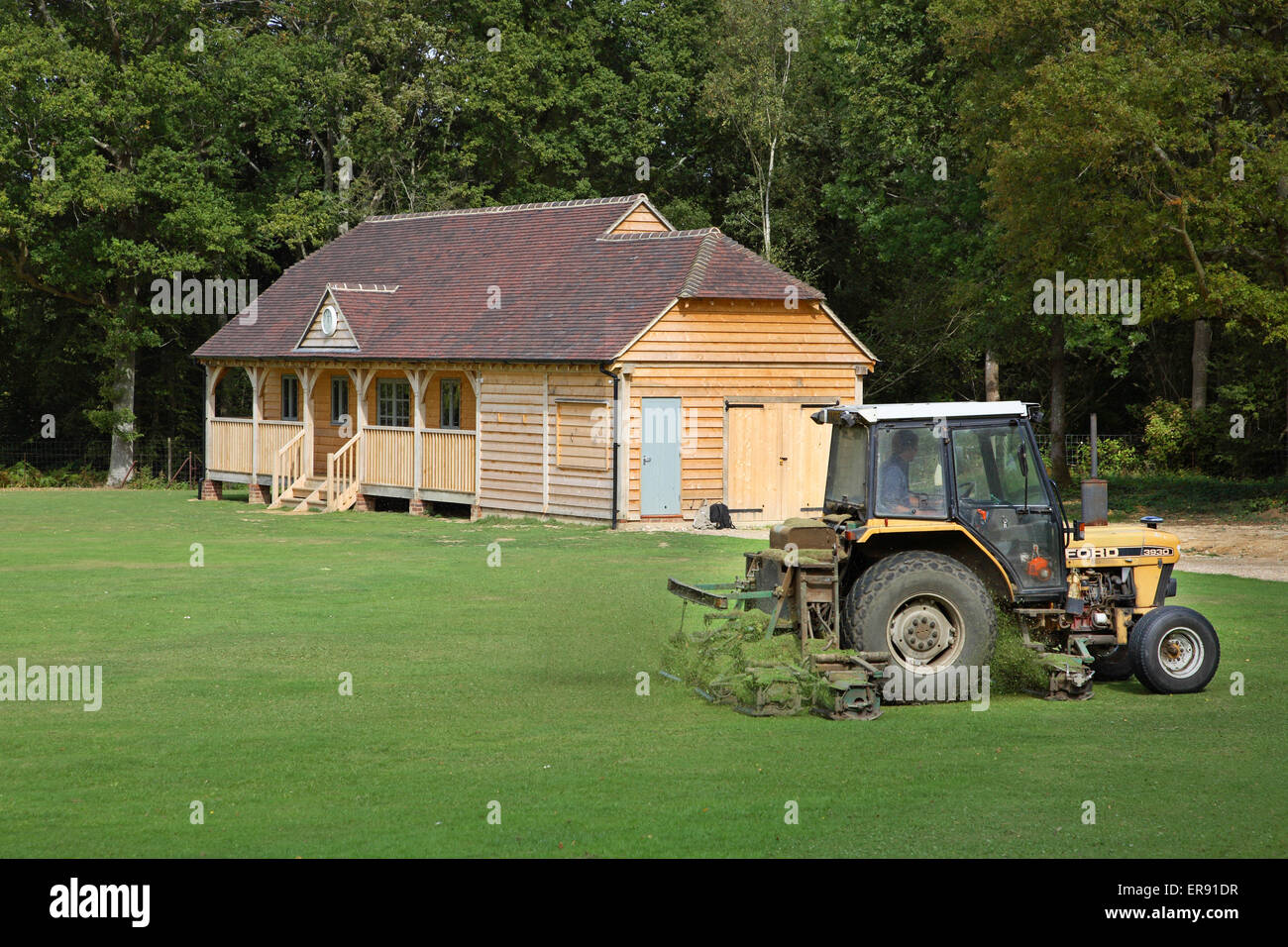 Un tracteur coupe de l'herbe en face d'un bois de construction traditionnelle construction pavillon cricket anglais dans le sud de l'Angleterre Banque D'Images