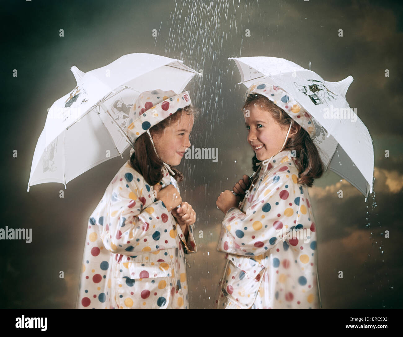 1960 SMILING JUMELLES portant des manteaux de pluie à pois ET DES CHAPEAUX D'AGIR AUX TERMES DE PARAPLUIES DANS LA PLUIE Banque D'Images