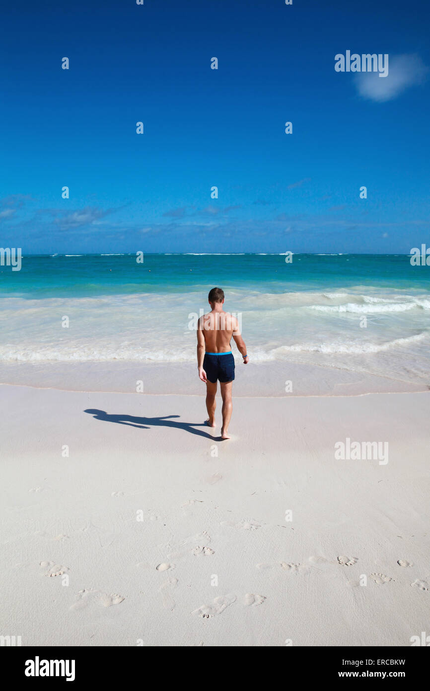 Jeune homme sportif va à l'océan sur une côte sableuse sous ciel bleu profond, vue arrière Banque D'Images