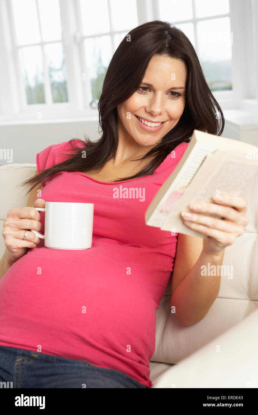 Femme enceinte de boire une boisson chaude et de la lecture livre à la maison Banque D'Images