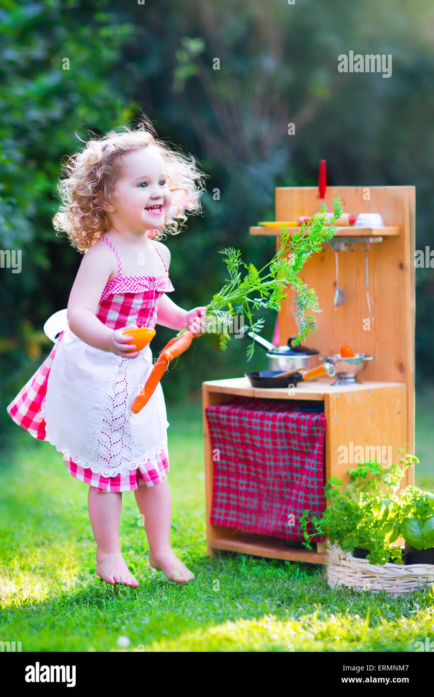 Curly Adorable Bebe Fille Portant Une Robe Rouge Et Un Tablier Blanc En Dentelle S Amusant A Jouer Avec Un Jouet Cuisine Poupee A Sunny Garden Photo Stock Alamy
