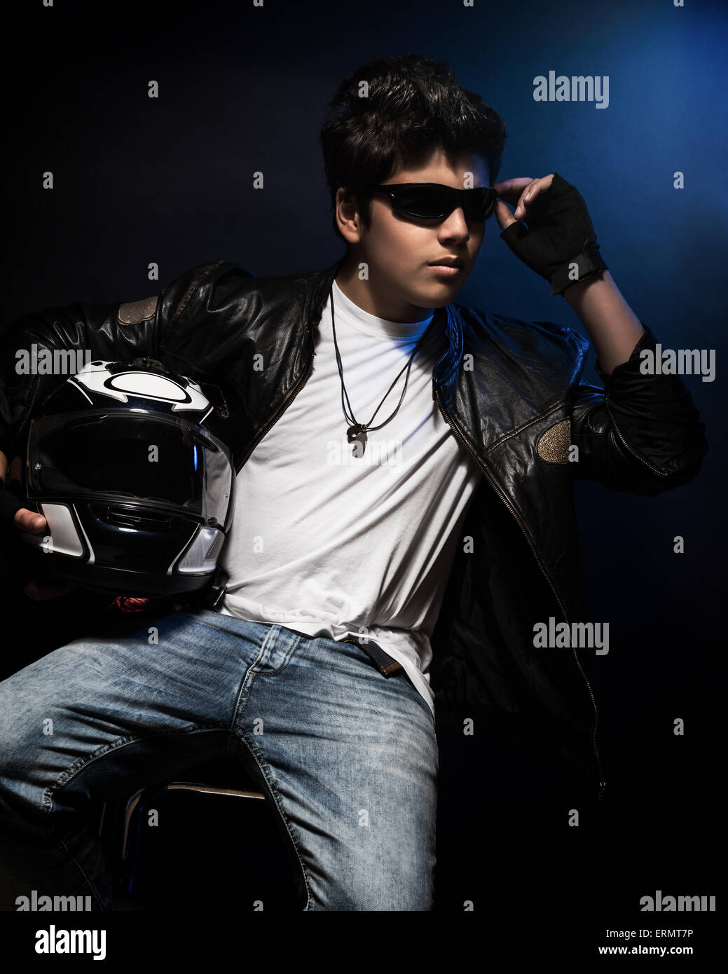Ado garçon élégant biker assis et posant dans le studio sur fond bleu foncé avec le casque, mains, mode pour motard Banque D'Images