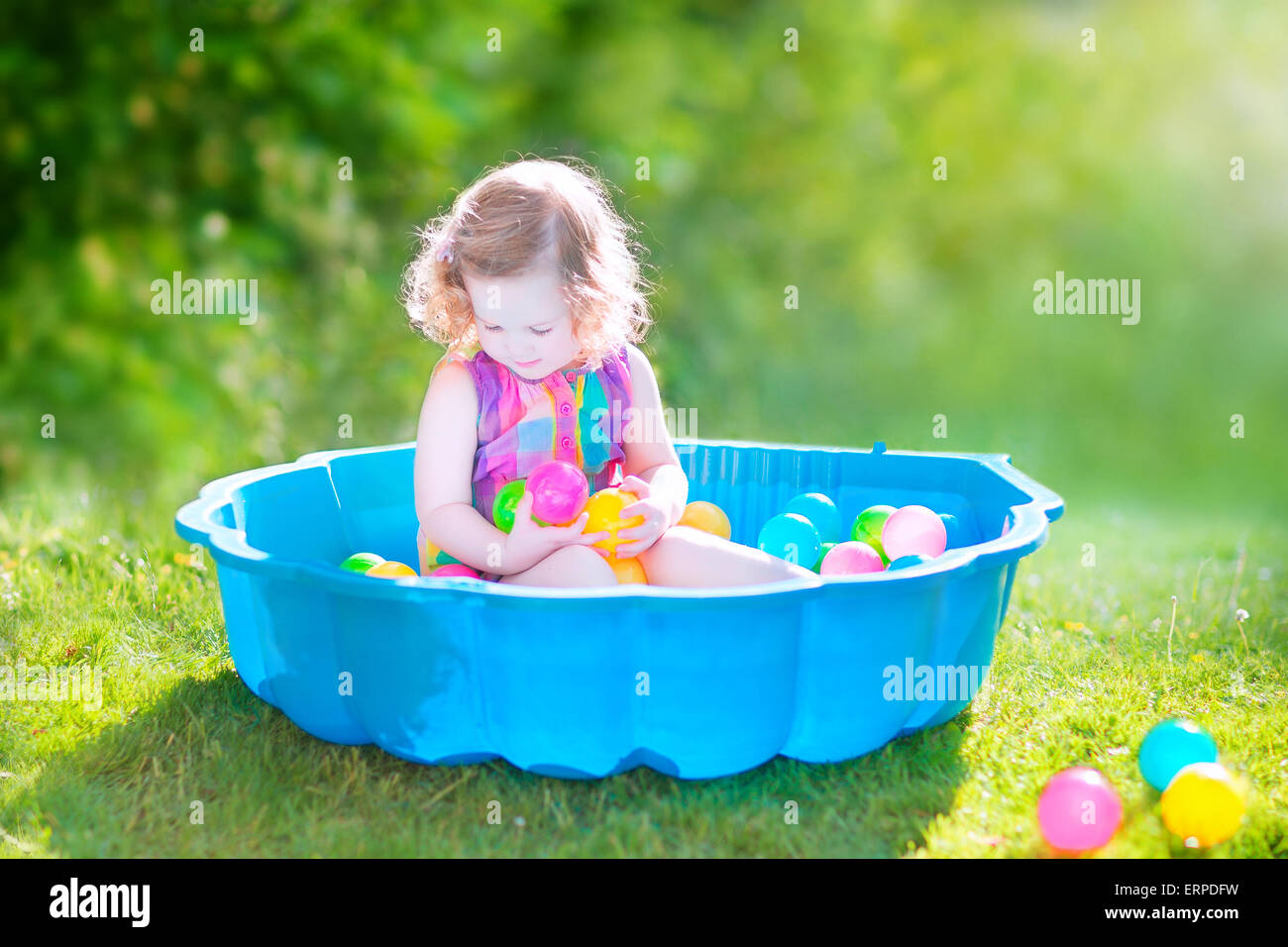 Happy cute girl avec des cheveux bouclés portant une robe colorée rose jouant dans un bac à sable avec des jouets en plastique billes dans jardin Banque D'Images