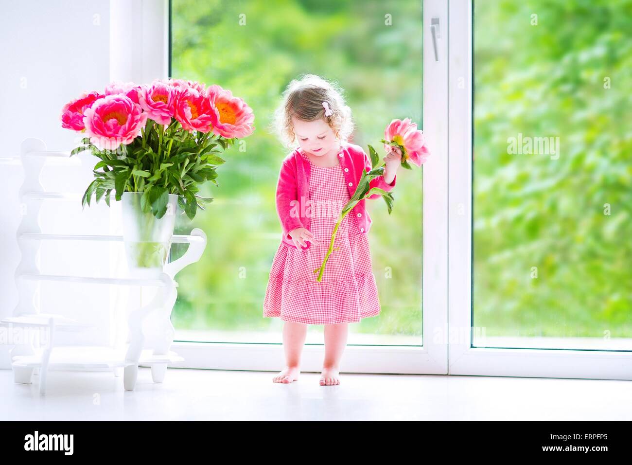 Heureux Mignon Bebe Fille Avec Des Cheveux Boucles Portant Une Robe Rose De Jouer Avec Un Tas De Belles Grosses Pivoines Dans Un Vase Photo Stock Alamy