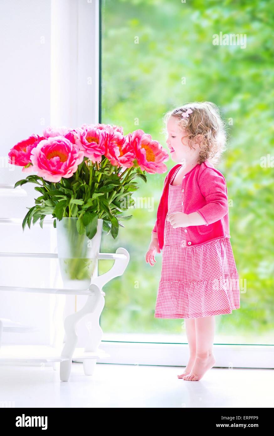 Heureux Mignon Bebe Fille Avec Des Cheveux Boucles Portant Une Robe Rose De Jouer Avec Un Tas De Belles Grosses Pivoines Dans Un Vase Photo Stock Alamy