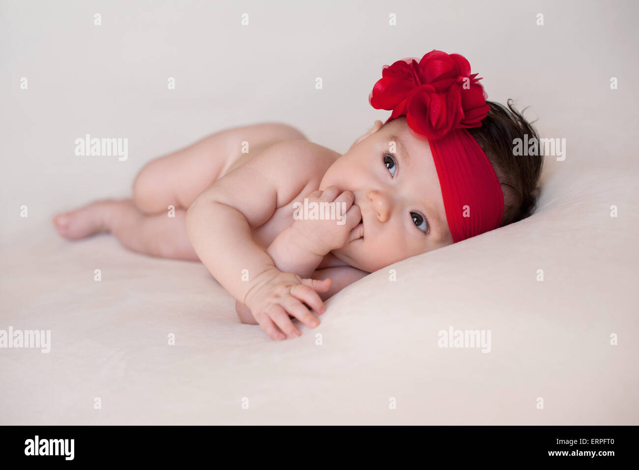 Un portrait d'une alerte, 3 mois baby girl wearing a grande, rouge, fleur serre-tête. Elle est couchée sur le côté, sur une couleur crème Banque D'Images