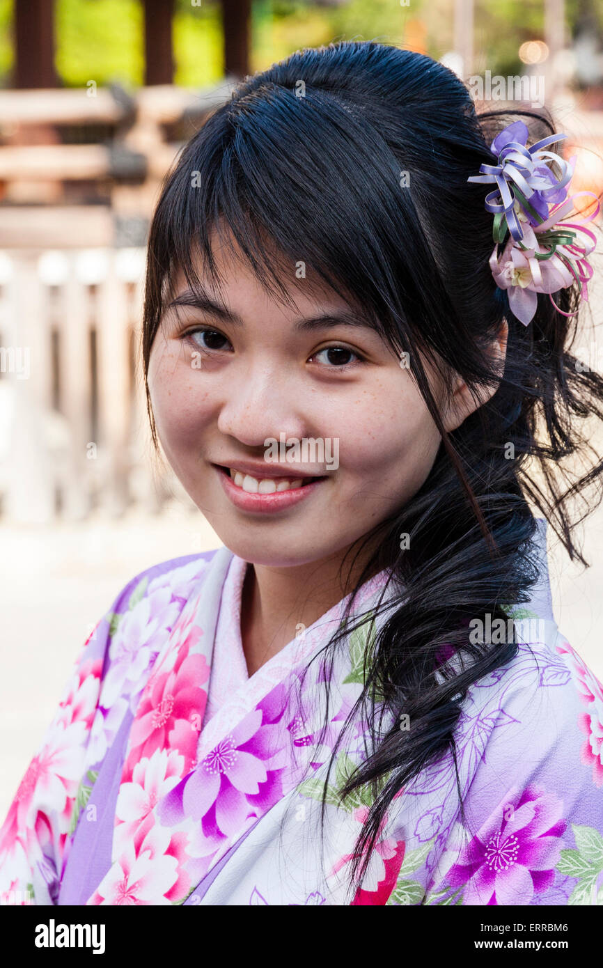 Tête et épaules une jeune femme chinoise souriant au spectateur. Elle porte un kimono violet et rose et a des fleurs dans ses cheveux. Sourire. Banque D'Images