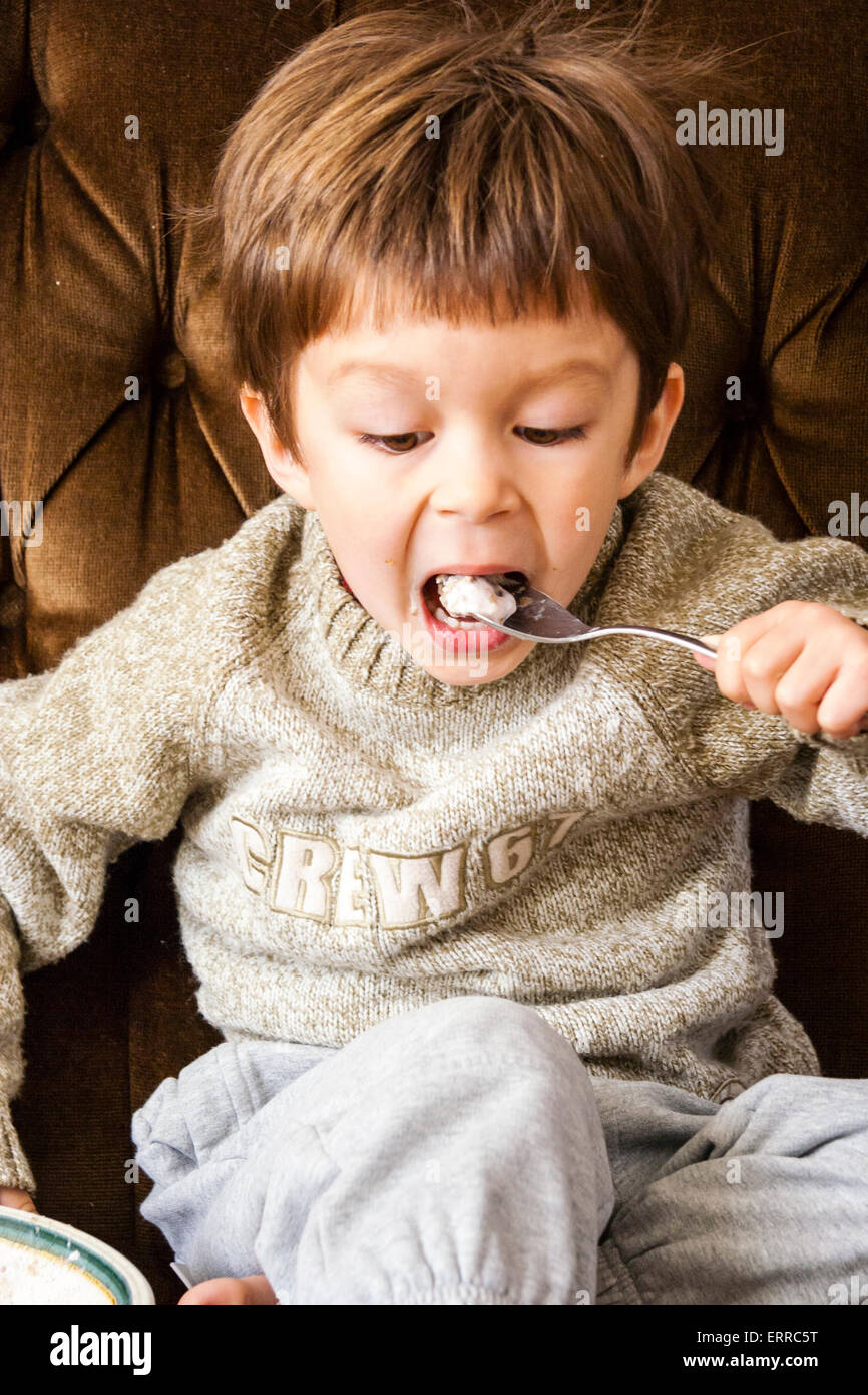 À l'intérieur, près d'un enfant caucasien, garçon de 3-4 ans, portant un épais cavalier et assis sur un settee brun tout en mangeant des céréales avec une cuillère d'argent. Banque D'Images