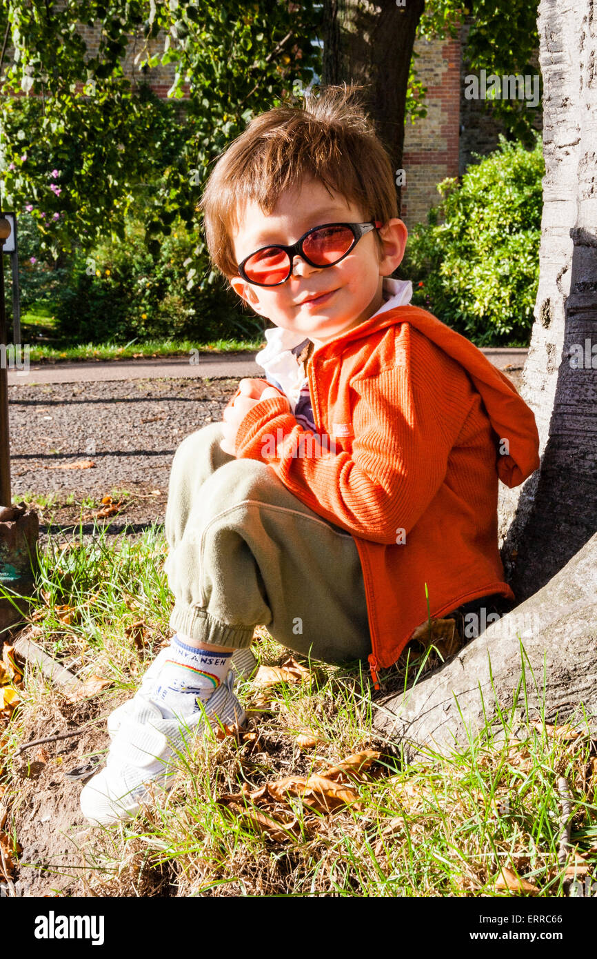 Enfant caucasien, garçon de 3-4 ans, assis à l'extérieur avec dos contre l' arbre et portant des lunettes de soleil. Visage tourné vers le spectateur.  Contact visuel, sourire Photo Stock - Alamy