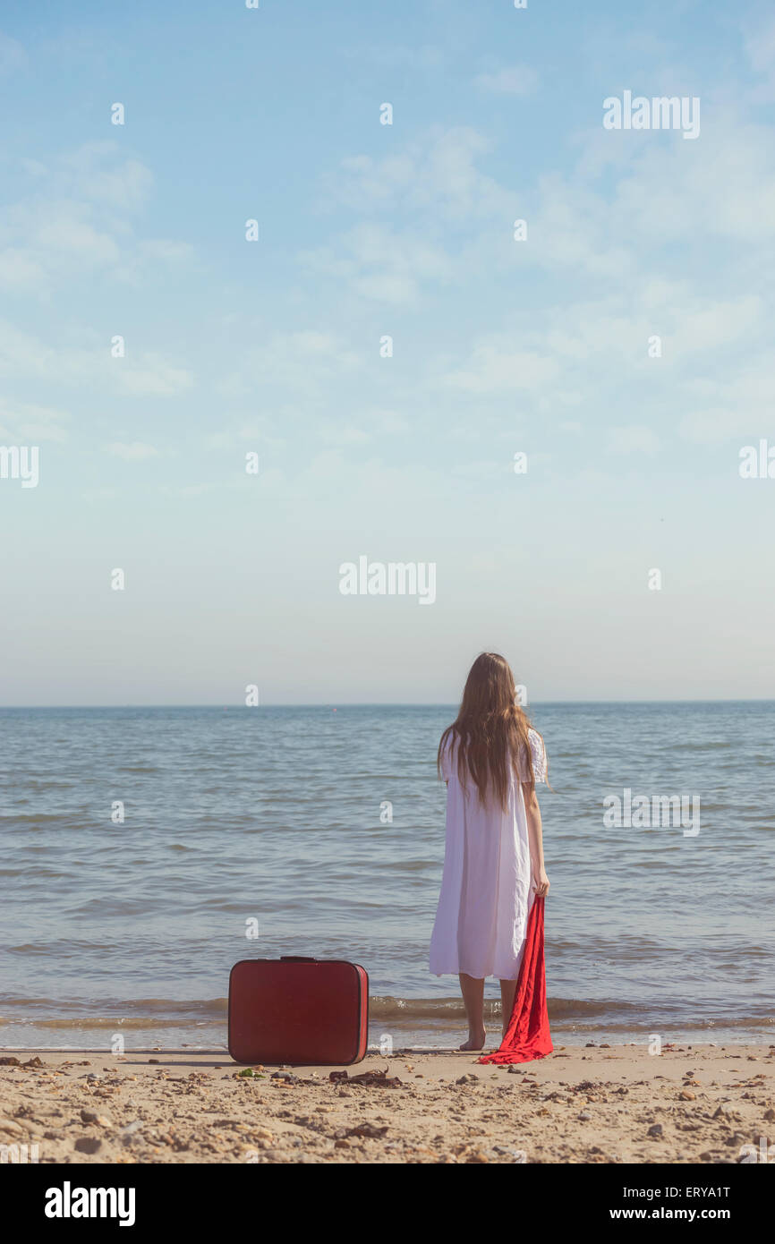 Une jeune fille en robe blanche est debout sur une plage avec une écharpe rouge et laissé une valise rouge derrière elle Banque D'Images