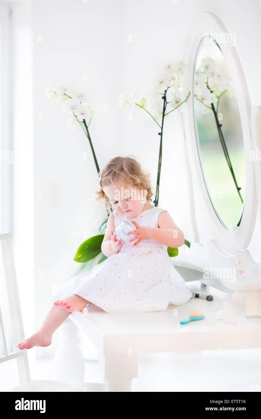 Heureux bébé fille avec des cheveux bouclés vêtue d'une robe blanche à  jouer avec le maquillage et les cosmétiques en face d'un miroir rond Photo  Stock - Alamy