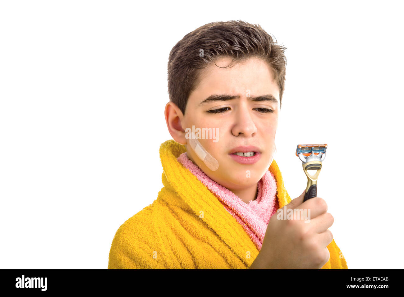 Un garçon de race blanche porte un peignoir jaune avec une serviette rose autour de son cou : il a des taches sur le visage et regarde tristement le rasoir qu'il a utilisé pour le rasage Banque D'Images
