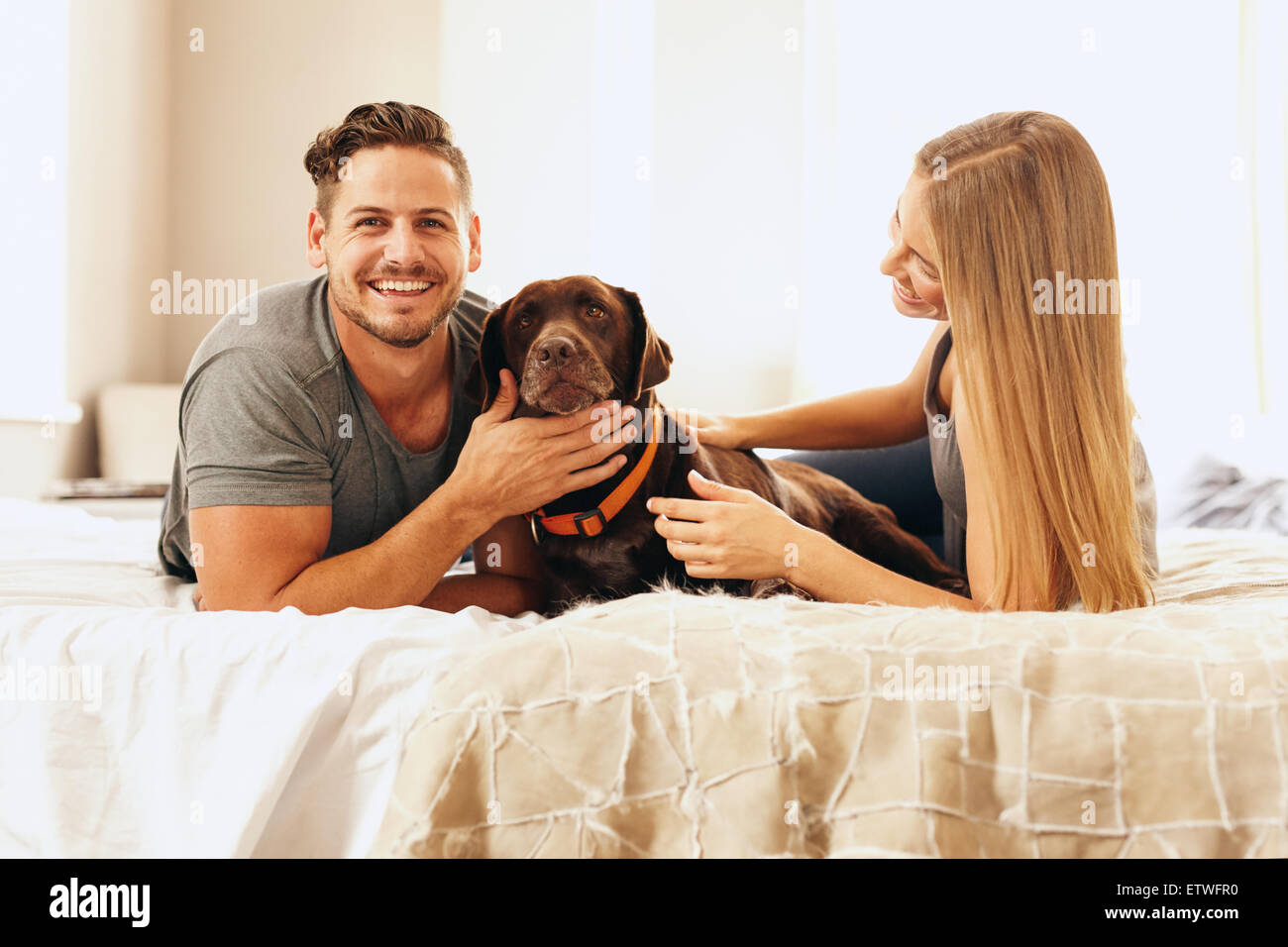Portrait of happy young couple on bed with dog in matin. L'homme de caresser le chien et souriant. Banque D'Images