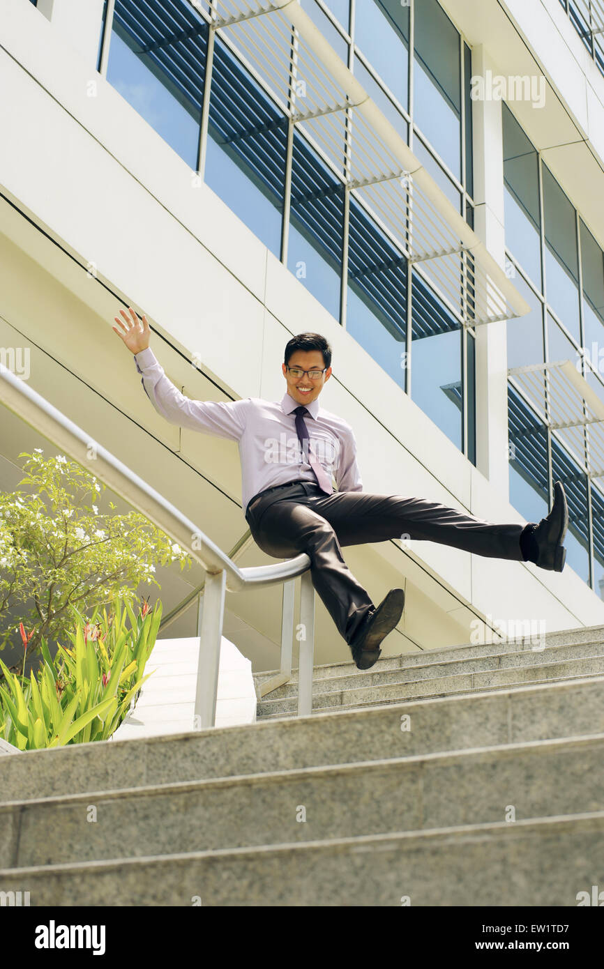 Jeune et heureux homme d'affaires chinois en bas coulissant sur des rails d'office d'escalier. L'homme apprécie sa nouvelle promotion à l'emploi Banque D'Images
