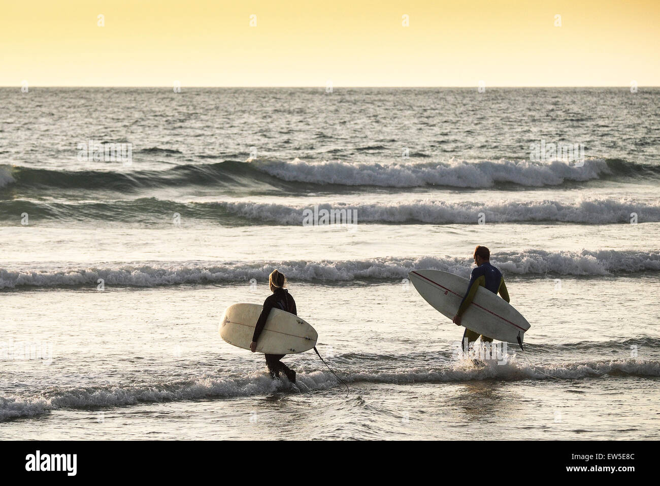 Deux surfeurs à pied dans la mer alors que le soleil se couche sur la plage de Fistral à Cornwall. UK. Banque D'Images