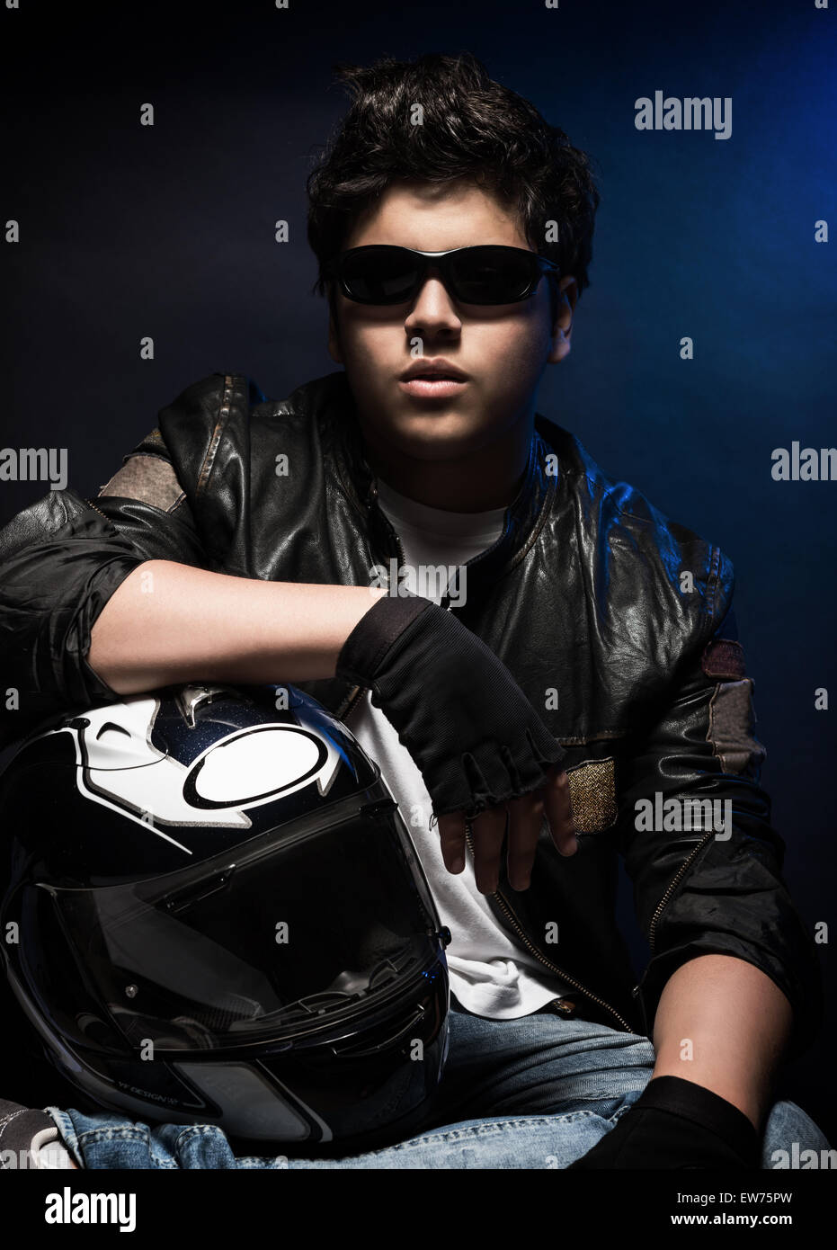 Portrait de joli garçon portant des lunettes de soleil, veste en cuir élégant et holding helmet posant dans le studio sur fond bleu Banque D'Images