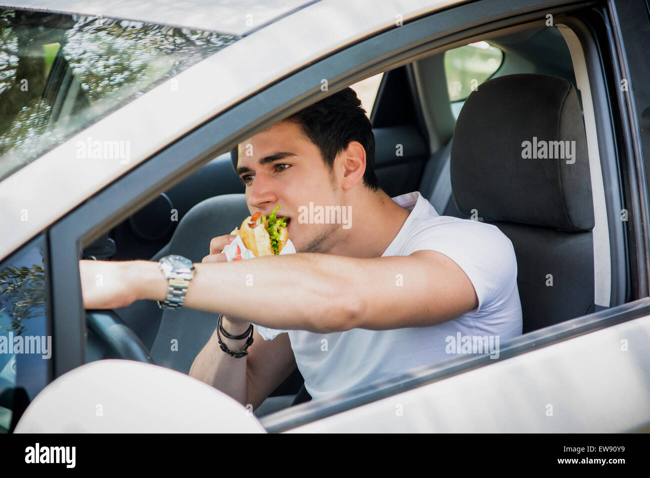 Beau jeune homme conduisant sa voiture tout en mangeant de la nourriture dans le trafic Banque D'Images