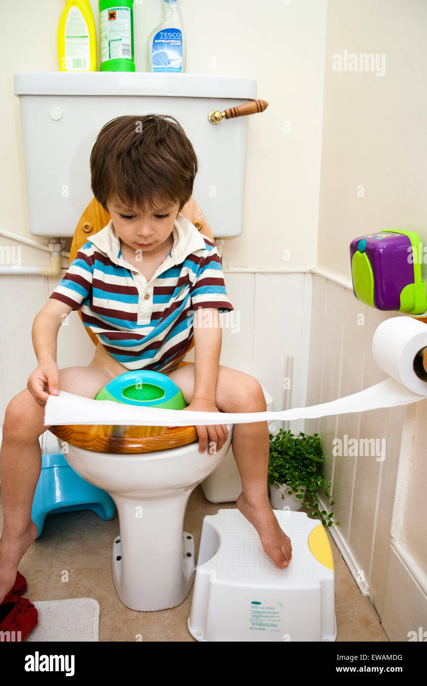 Caucasien enfant, garçon, 4-5 ans, en face, assis sur un siège de toilette  enfant toilettes et en tirant sur le papier toilette du support sur le mur  à côté de lui Photo