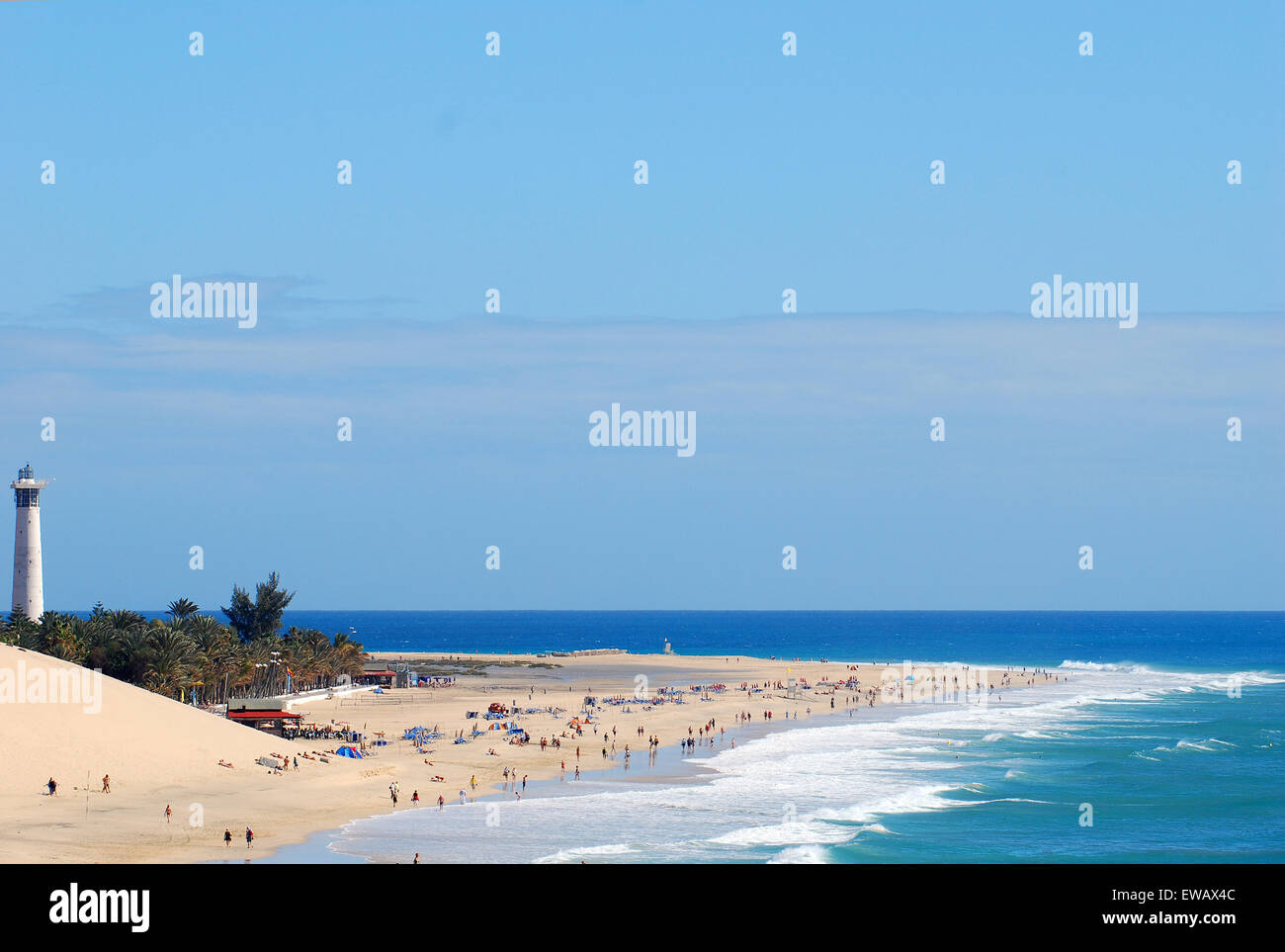 Une plage de sable ensoleillée être frappé doucement en crêtes de vagues écumeuses, avec un phare debout sur une pochette d'arbres tropicaux. Banque D'Images