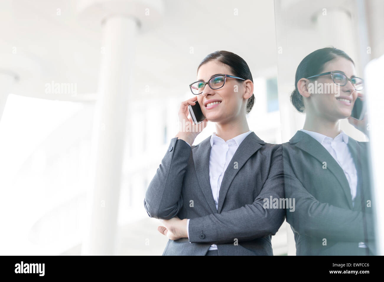 Happy businesswoman using cell phone while leaning on glass wall Banque D'Images