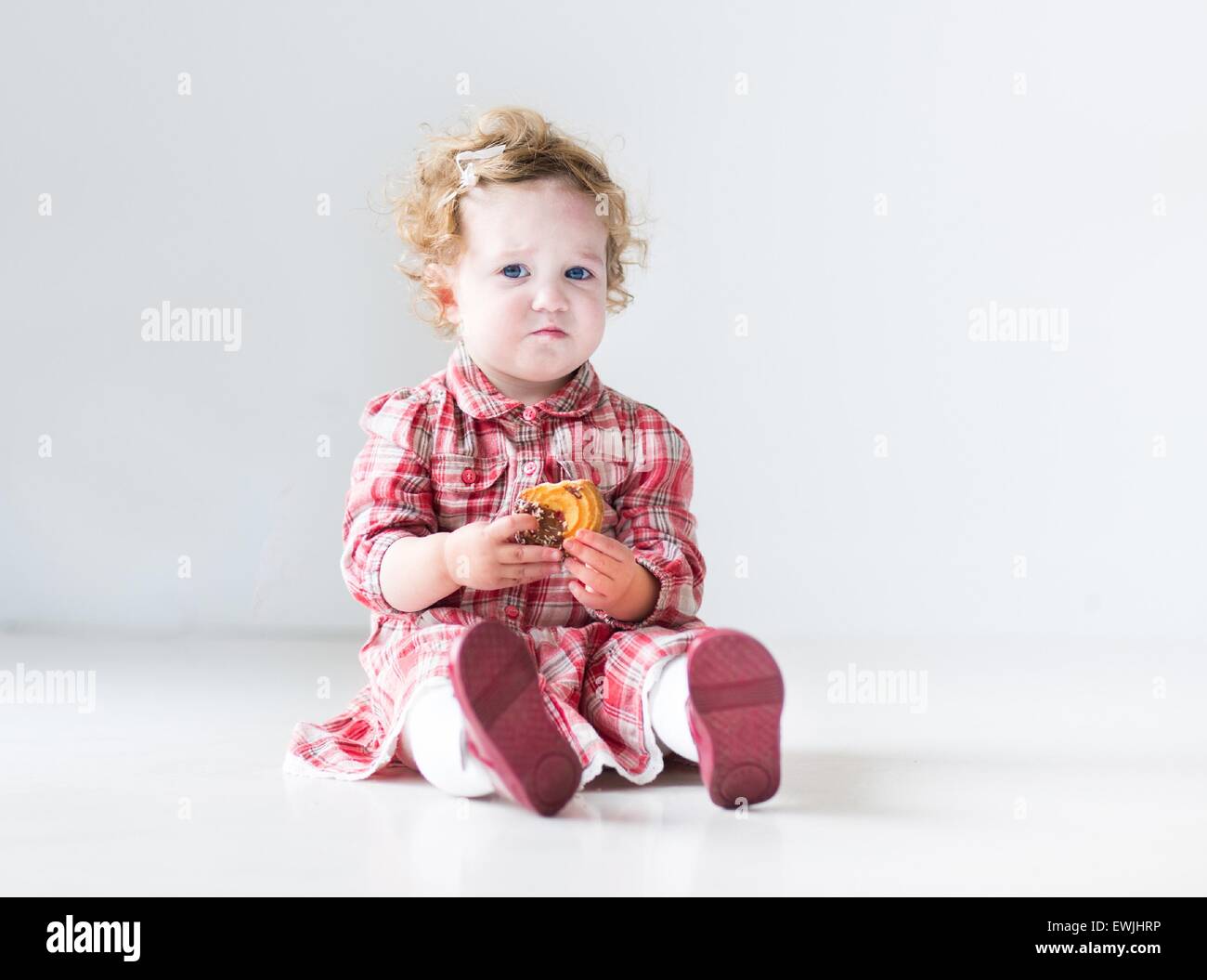 Funny baby girl avec des cheveux bouclés portant une robe rouge de manger un biscuit de Noël dans une pépinière blanc Banque D'Images