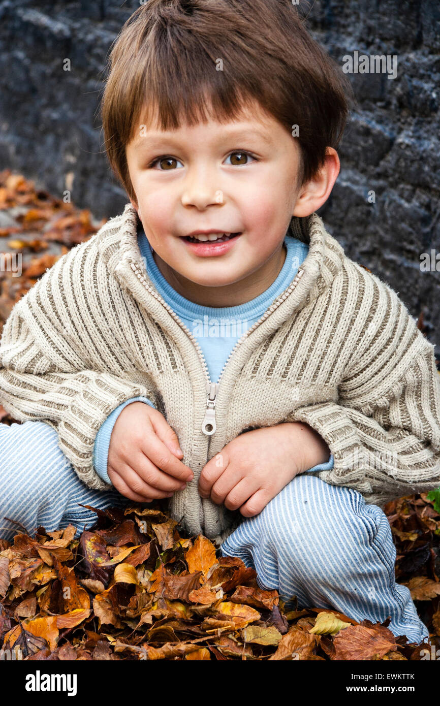 Caucasien enfant, garçon, 5-6 ans, à l'extérieur par un mur de brique peint en noir, accroupi entre brown l'automne les feuilles tombées, looking up and smiling. Banque D'Images
