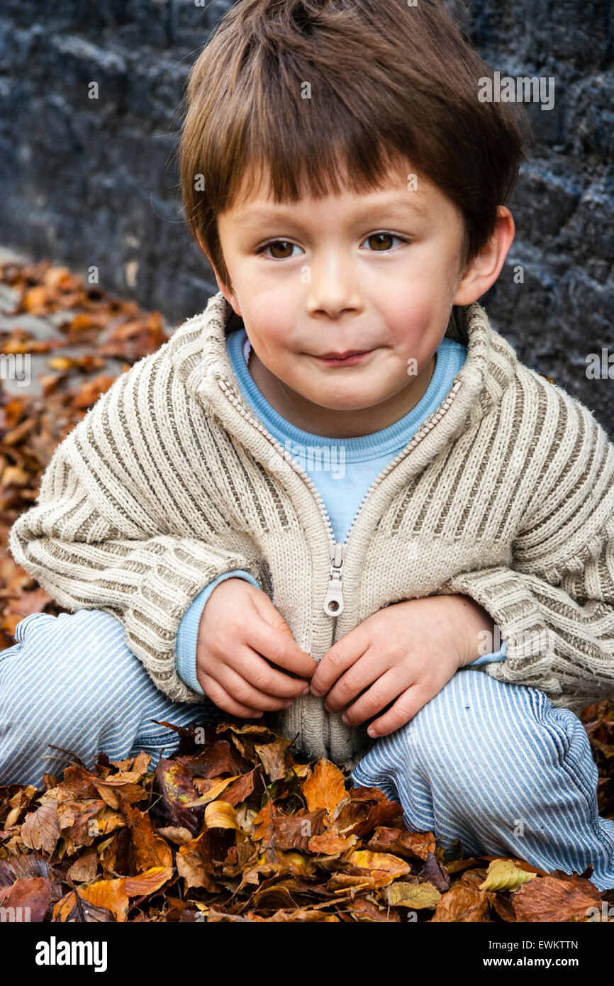 Caucasien enfant, garçon, 5-6 ans, à l'extérieur par un mur de brique peint en noir, accroupi entre brown automne les feuilles tombées, regardant vers le bas de façon réfléchie. Banque D'Images
