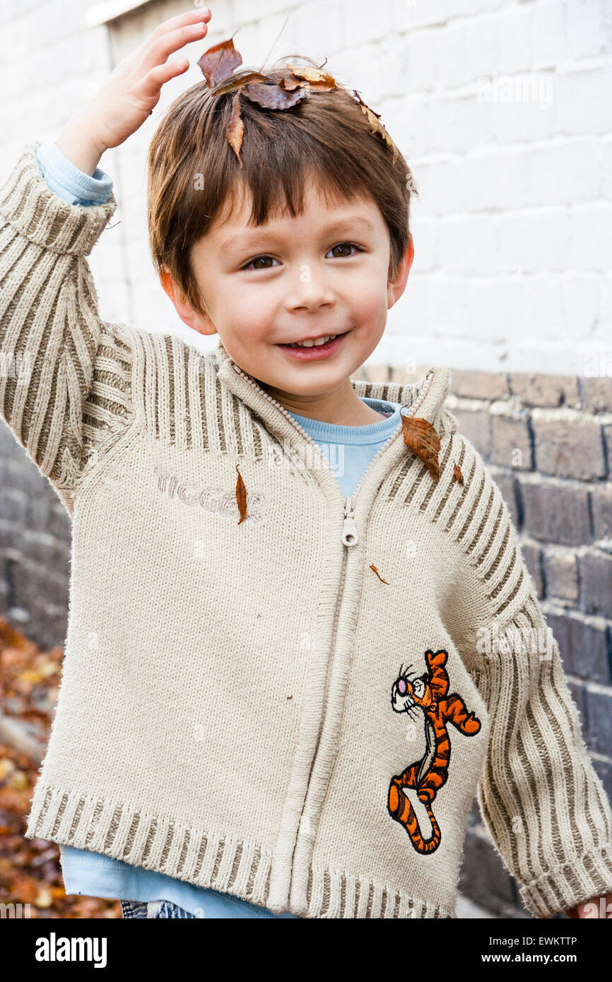Close up of caucasien enfant, garçon, 5-6 ans, à l'extérieur par un blanc et mur de briques peint en noir, mettre les feuilles d'automne marron sur sa tête, tout en souriant. Banque D'Images