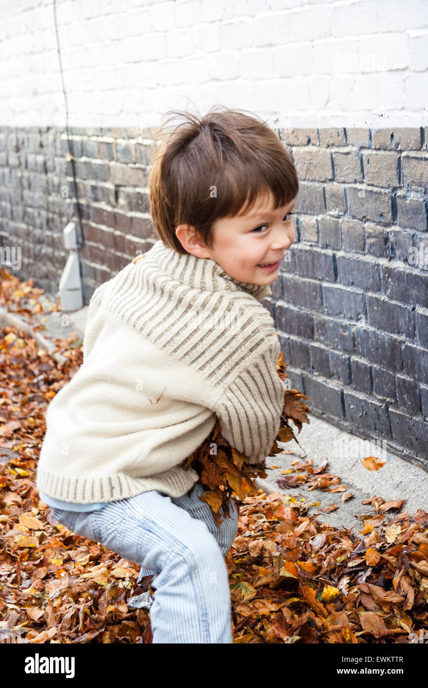 Close up of caucasien enfant, garçon, 5-6 ans, à l'extérieur en rue par un mur de brique peint, tournant et sur le point de jeter brassée de feuilles d'automne au spectateur. Banque D'Images