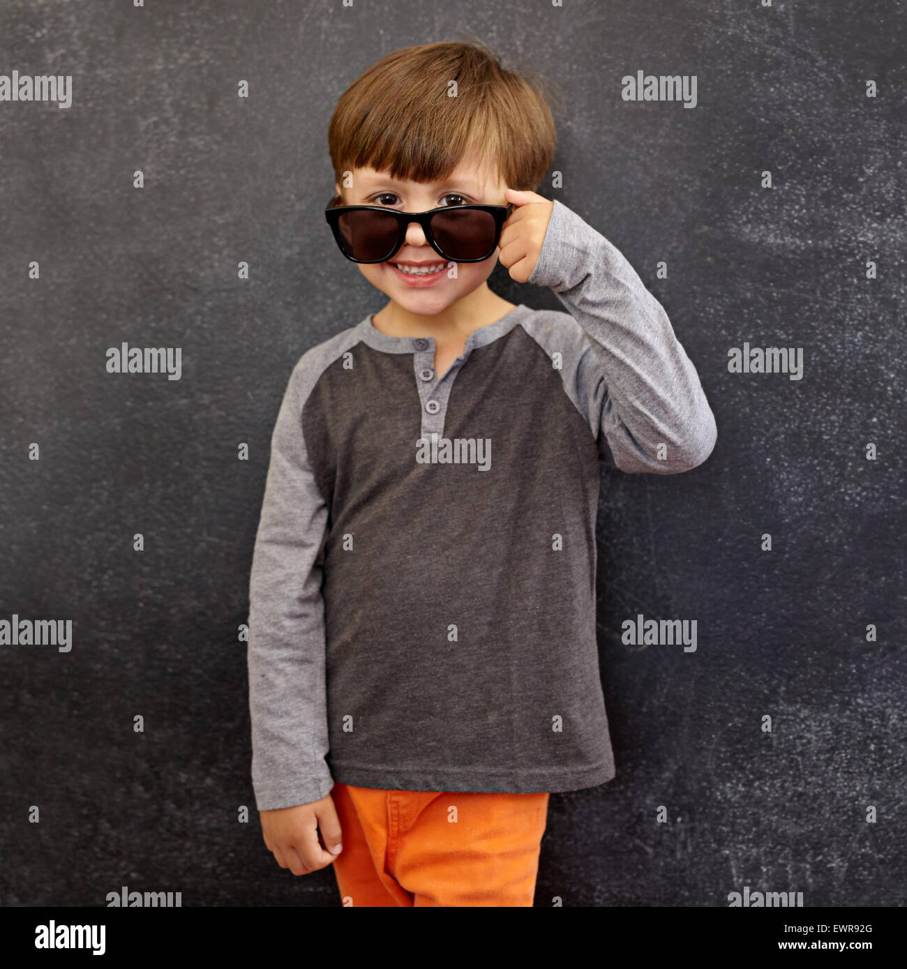 Portrait of happy little boy wearing sunglasses smiling at camera. Petit boy peering over ses lunettes contre tableau noir. Banque D'Images
