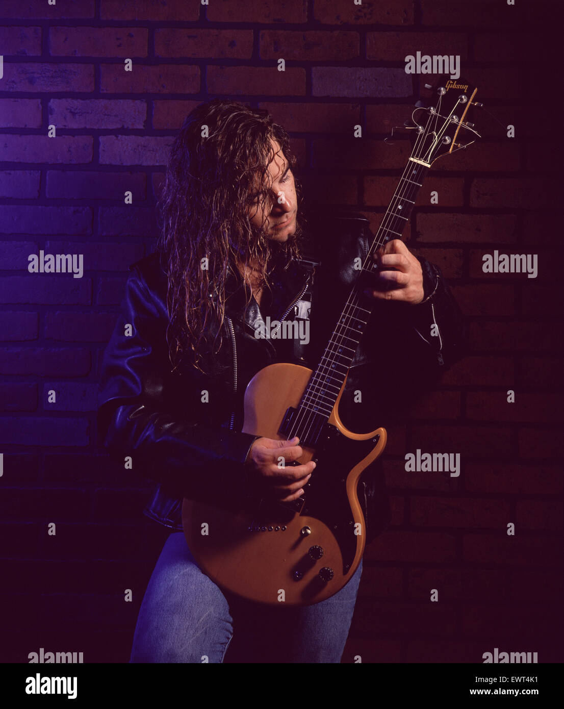 Guitariste rock avec de longs cheveux blonds, portant une veste en cuir, jouant de la guitare électrique contre un mur de briques. Banque D'Images
