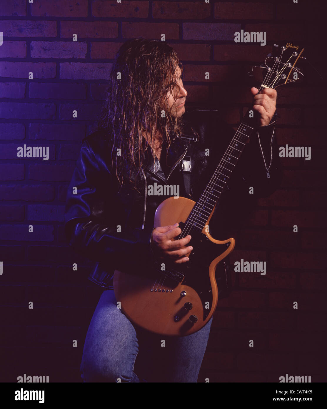 Guitariste rock avec de longs cheveux blonds, portant une veste en cuir, jouant de la guitare électrique contre un mur de briques. Banque D'Images