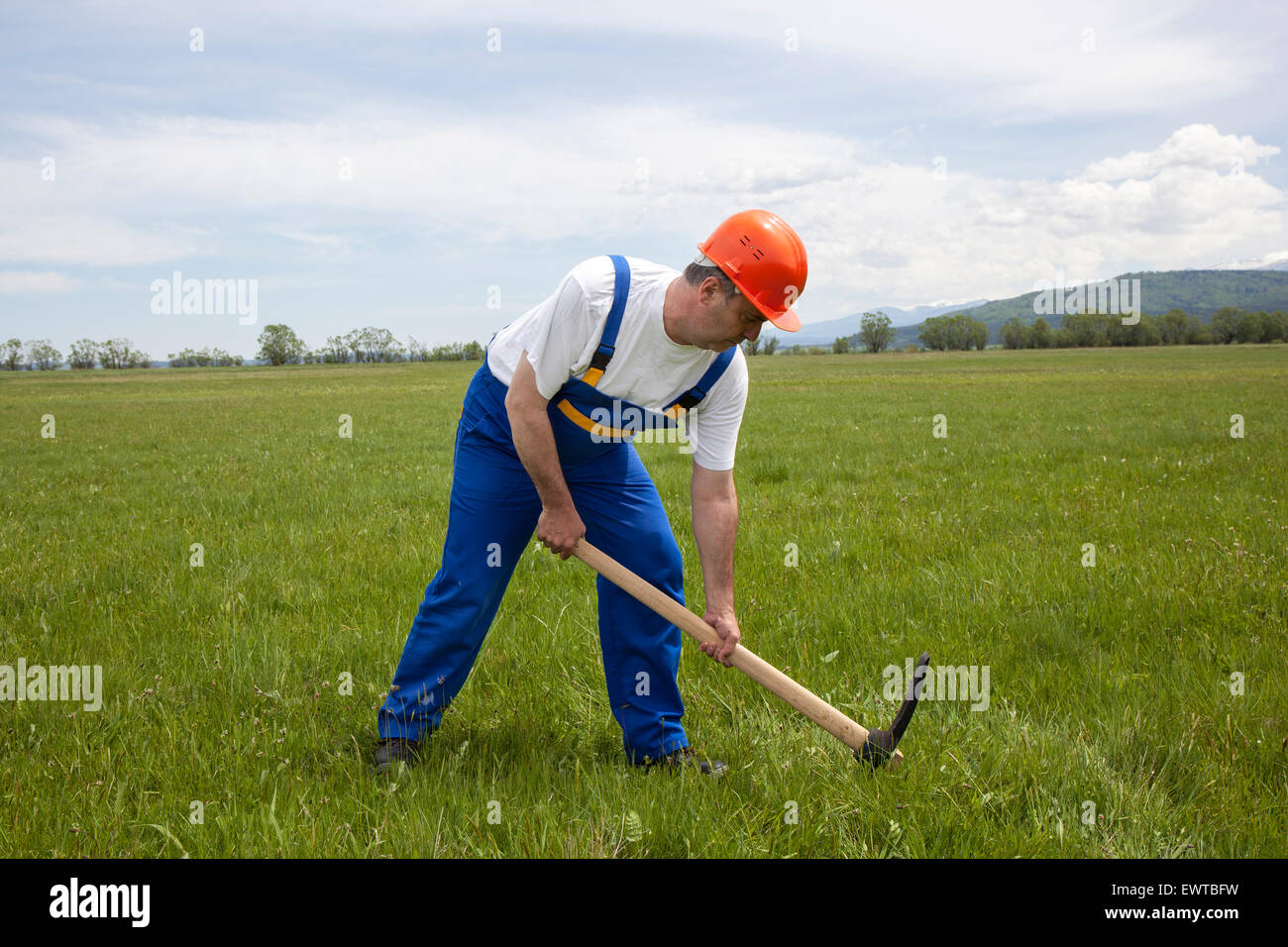 Image d'un travailleur dans les vêtements de travail et casque creuser avec une pioche sur le champ vert. Banque D'Images