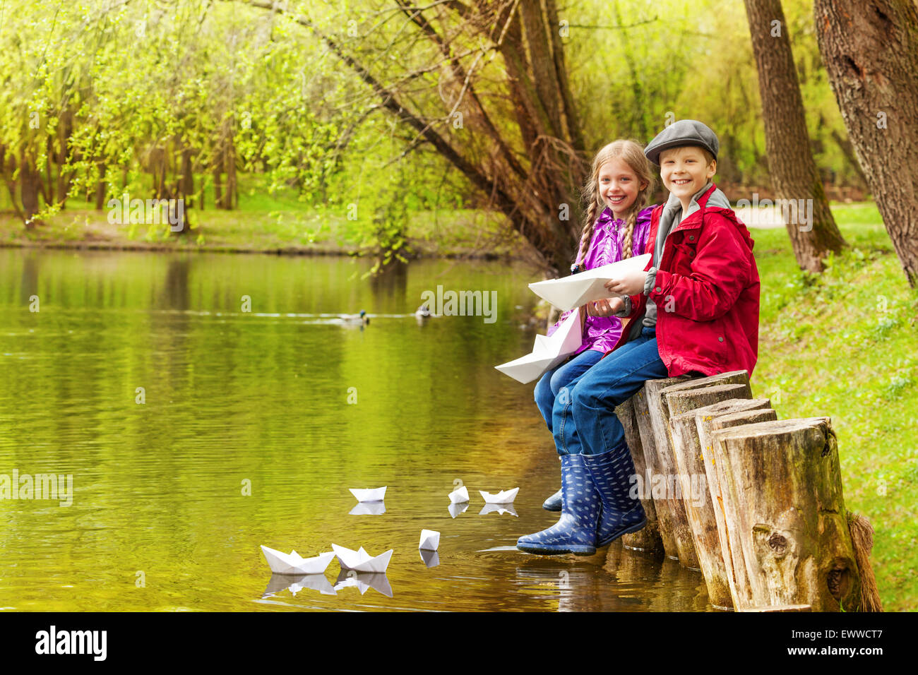 Les enfants assis près de l'étang jouer avec bateaux en papier Banque D'Images