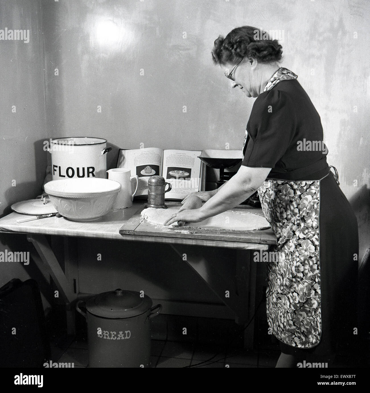 Années 1950, historique, sur une petite table de marbre dans un coin d'une pièce, une dame d'âge mûr portant un tablier, faire de la pâtisserie, à l'aide d'un rouleau à pâtisserie en bois sur une planche pour l'aplatir et façonner l'dougn avant la cuisson. Banque D'Images